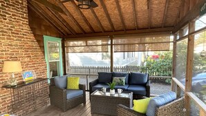 Un porche cubierto con muebles de mimbre, una mesa con tazas y una planta, y vista a una cerca blanca de piquetes.