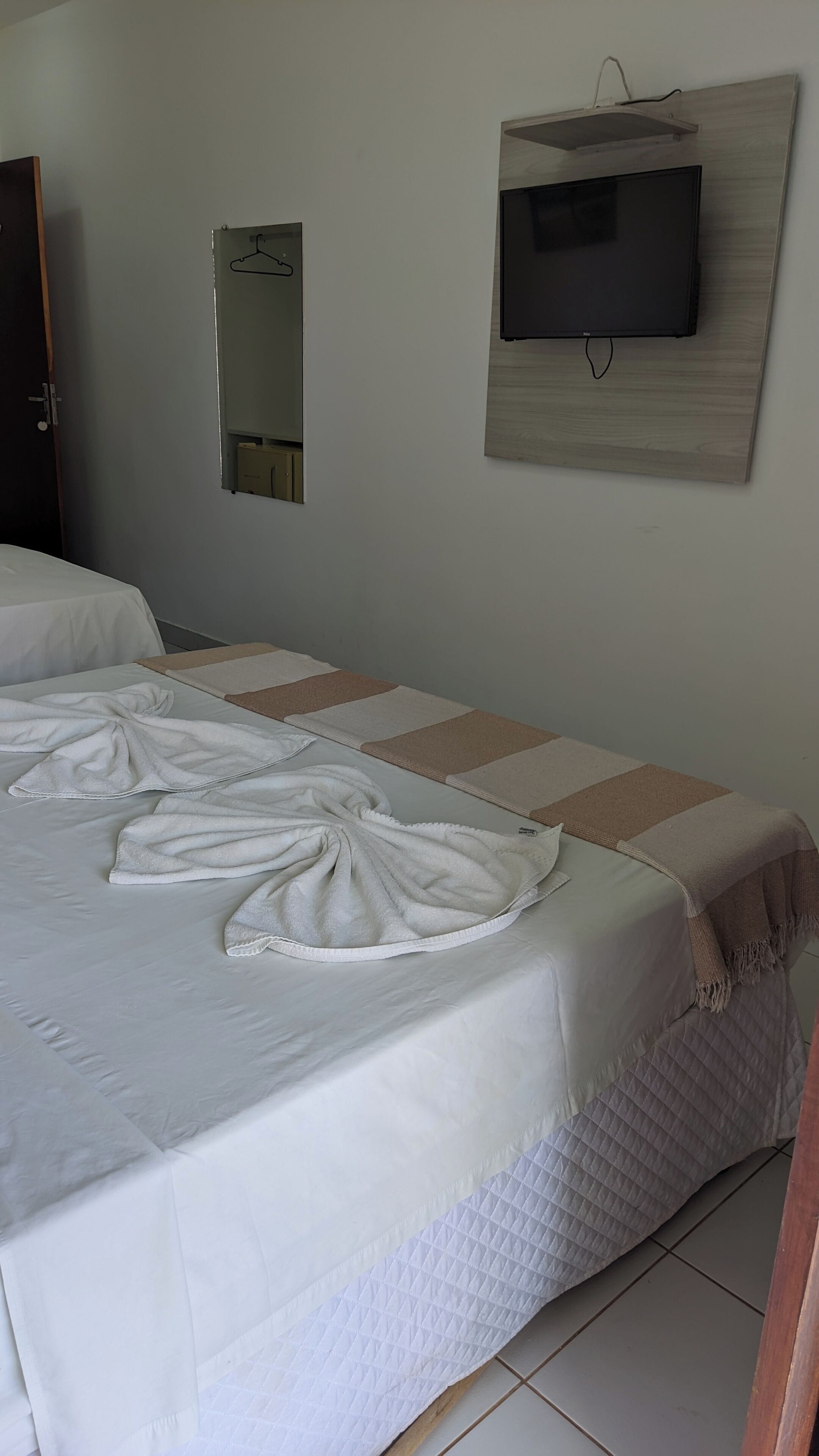 A neatly made bed with towels, a television mounted on the wall, and a mirror above it.