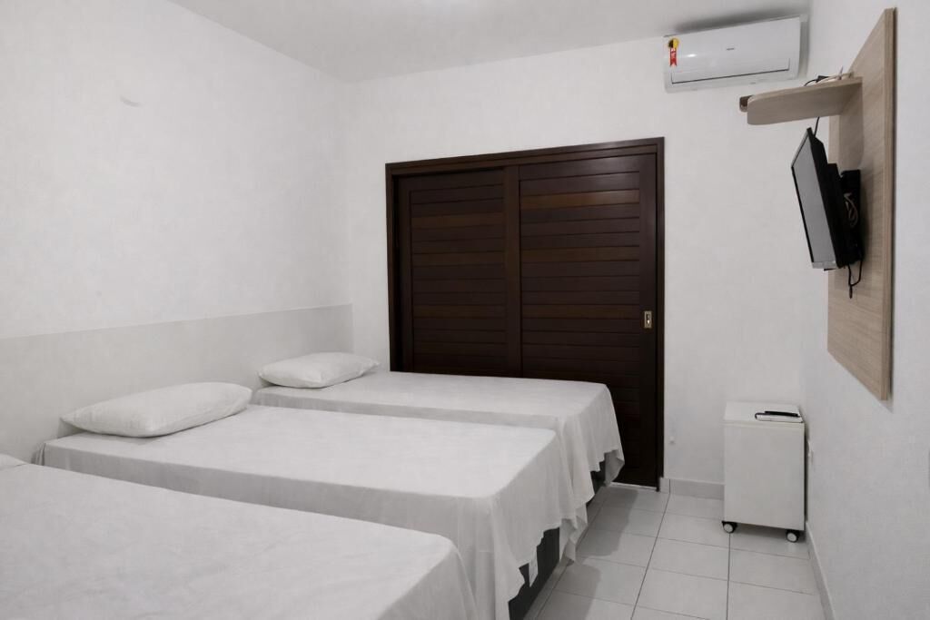 Quarto com duas camas de solteiro, um guarda-roupa de madeira, uma pequena lixeira branca e uma televisão montada na parede.