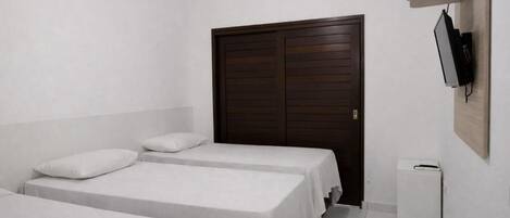 Quarto com duas camas de solteiro, um guarda-roupa de madeira, uma pequena lixeira branca e uma televisão montada na parede.