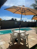 Zona de piscina al aire libre con mesa y sillas, rodeada de sombrillas y un patio empedrado.
