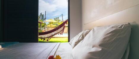 A hotel room with a bed, a view of a hammock outside, and a window.