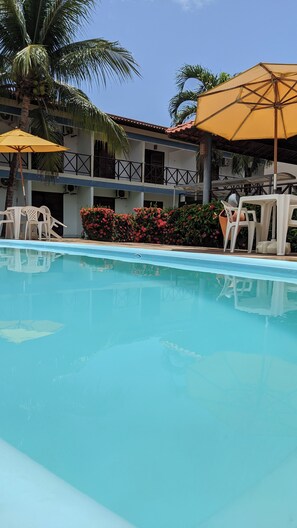 A swimming pool with white lounge chairs and yellow umbrellas, surrounded by a building with balconies and a palm tree. - Pousada Solar da Praia (Tamandaré)