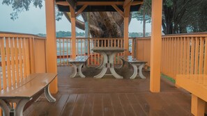 An outdoor wooden deck with a central table and benches, surrounded by orange railings and a large tree in the background.