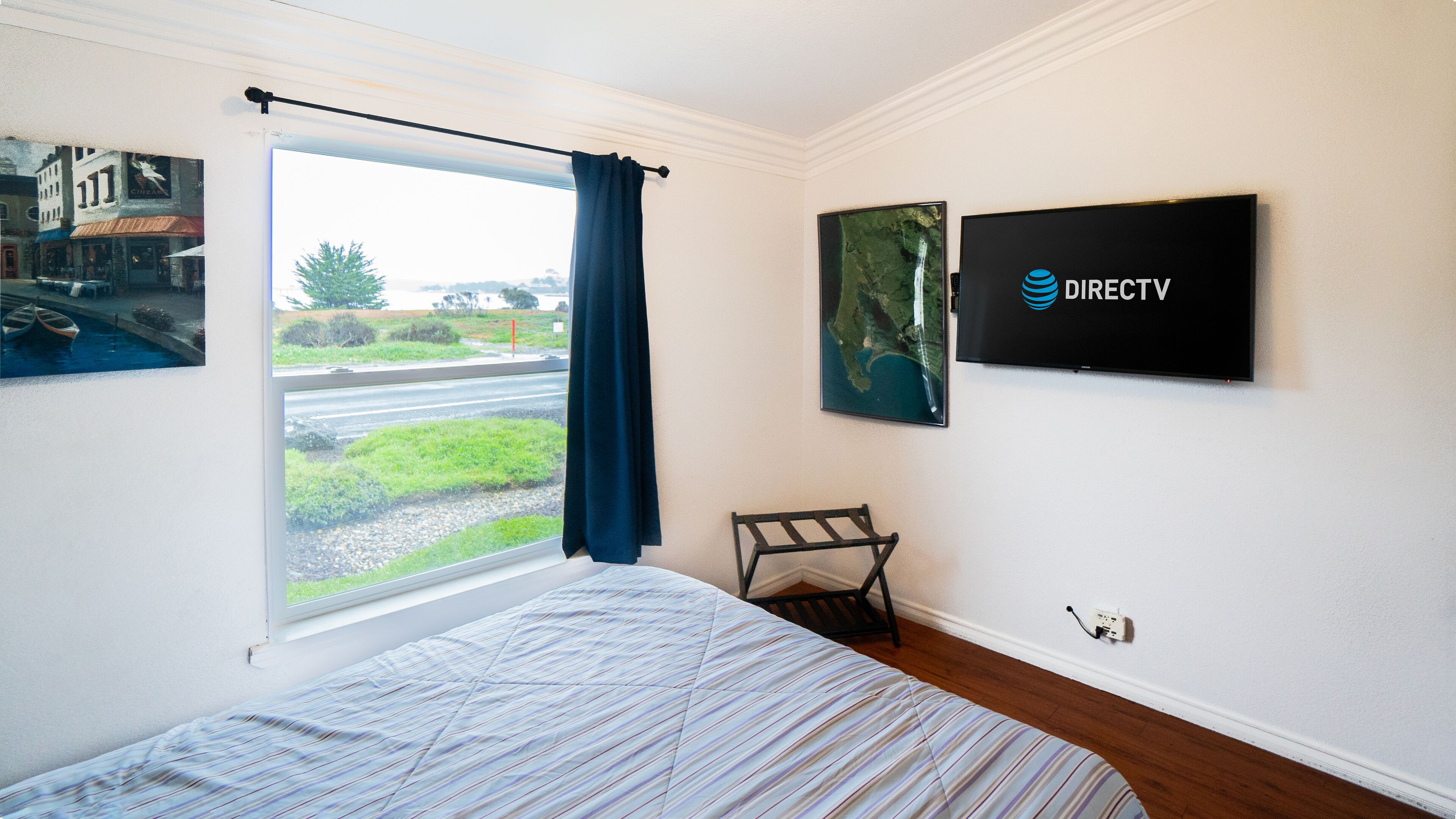A hotel room with a bed, a window with curtains, a chair, a wall-mounted TV, and a framed picture.