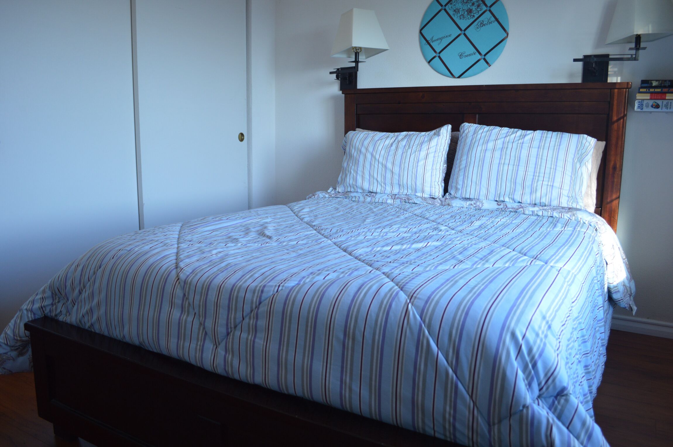 A neatly made bed with striped bedding, a wooden headboard, and a wall-mounted lamp.