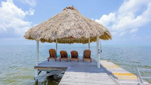 A thatched-roof gazebo with four wooden chairs on a wooden pier extending into calm waters. - An unBELIZEable home...Barefoot beachfront luxury! (San Pedro)