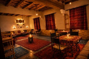 A cozy, dimly lit room with wooden beams, a sofa, a dining table with chairs, and a patterned rug. - Auberge La fibule du dades (Ait Sedrate Jbel El Soufla)