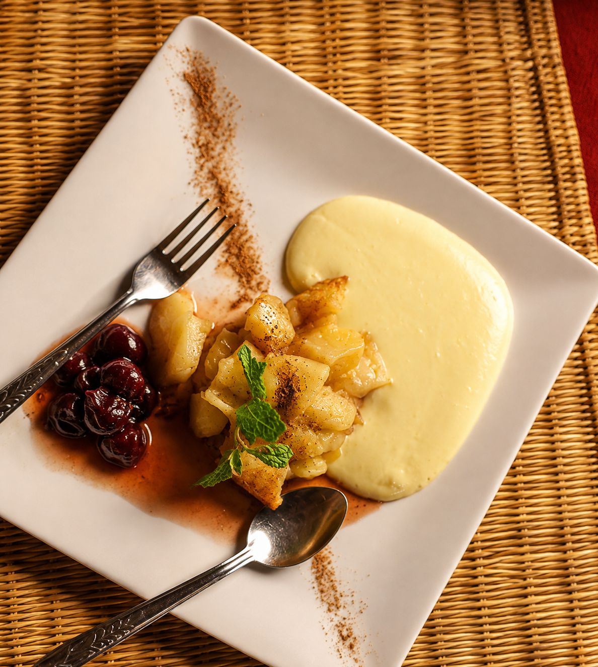 A square plate with a dessert consisting of diced fruit, a dollop of cream, and a mint garnish.