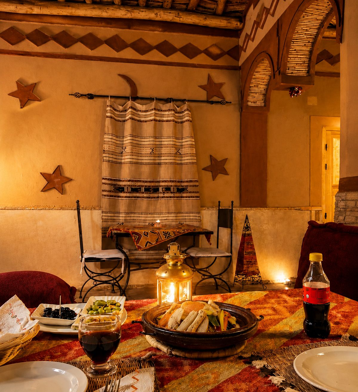 A dining area with a table set for two, featuring a traditional Moroccan tagine, a bottle of Coca-Cola, and a glass of red wine.