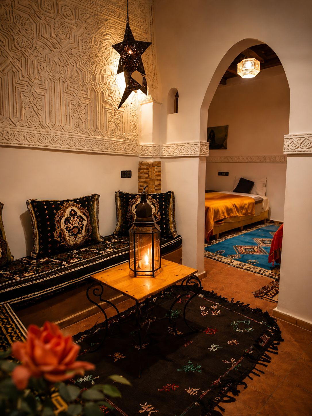 Une pièce chaleureuse avec un mur orné d’un motif, une suspension en forme d’étoile, une table en bois avec une lanterne, un canapé avec des coussins et un lit recouvert d’un tapis bleu.