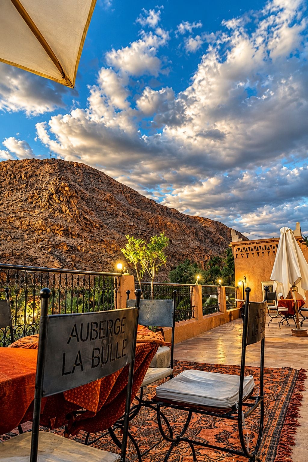 Un área de comedor al aire libre con mesas y sillas, un cartel que dice 'AUBERGE LA BOULE' y un telón de fondo montañoso.