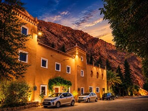 Et hotel med et bjerg i baggrunden, flere biler parkeret foran og en veloplyst indgang.