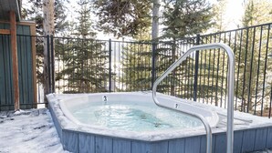 Un bain à remous entouré d’une clôture métallique, avec une terrasse en bois et de la neige au sol.