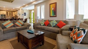 Ein Wohnzimmer mit einem Ecksofa, einem Couchtisch und farbenfrohen Kissen. An der Wand hängt ein Gemälde und ein Fenster mit Jalousien.