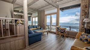 Ein großzügiger Wohnbereich mit einem großen Fenster und Meerblick. Es gibt ein blaues Sofa, einen Holztisch und einen Korbstuhl.