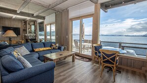 Ein großzügiger Wohnbereich mit einer großen, blauen Couch, einem hölzernen Couchtisch und Korbstühlen. Ein großes Fenster bietet Blick auf das Wasser und die Berge.