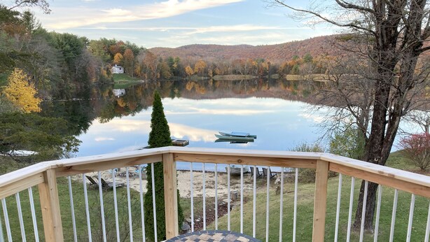 A deck with a round table and two chairs, overlooking a lake with a boat and a house in the background.