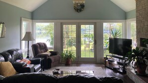 A spacious living room with a ceiling fan, a fireplace, a television, and a large window offering a view of a garden.