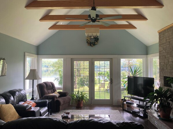 A spacious living room with a ceiling fan, a fireplace, a television, and a large window offering a view of a garden.