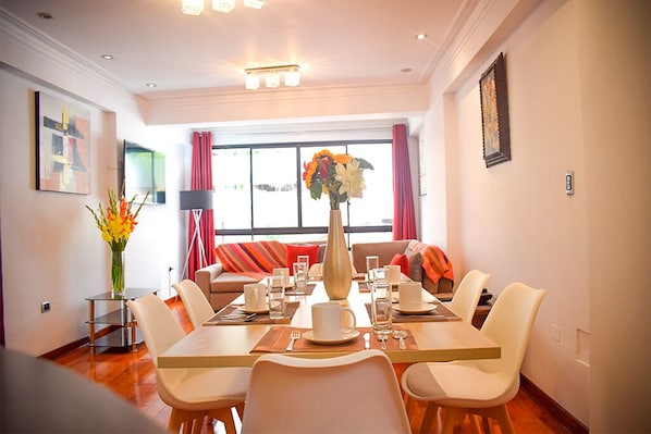 A dining area with a table set for four, a vase of flowers, and a sofa with red cushions.