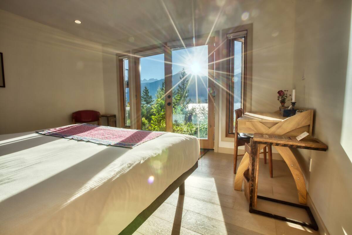 Una habitación con cama, una mesa de madera y vistas a montañas a través de amplios ventanales.