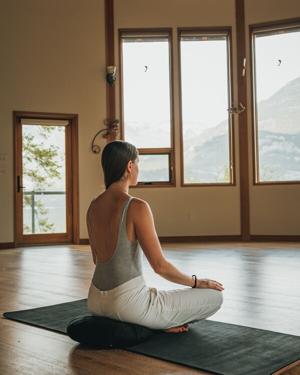 A person sitting on a yoga mat in a room with large windows and wooden floors. - The Sentinel (Kaslo)