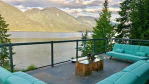 A balcony with turquoise cushioned seating, a wooden coffee table, and a view of a lake with mountains in the background. - The Sentinel (Kaslo)