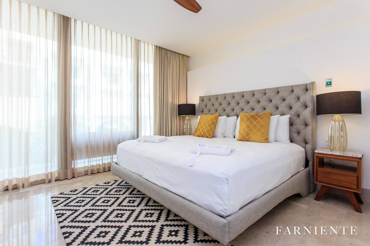 A hotel room with a large bed, a patterned rug, a nightstand with a lamp, and sheer curtains.