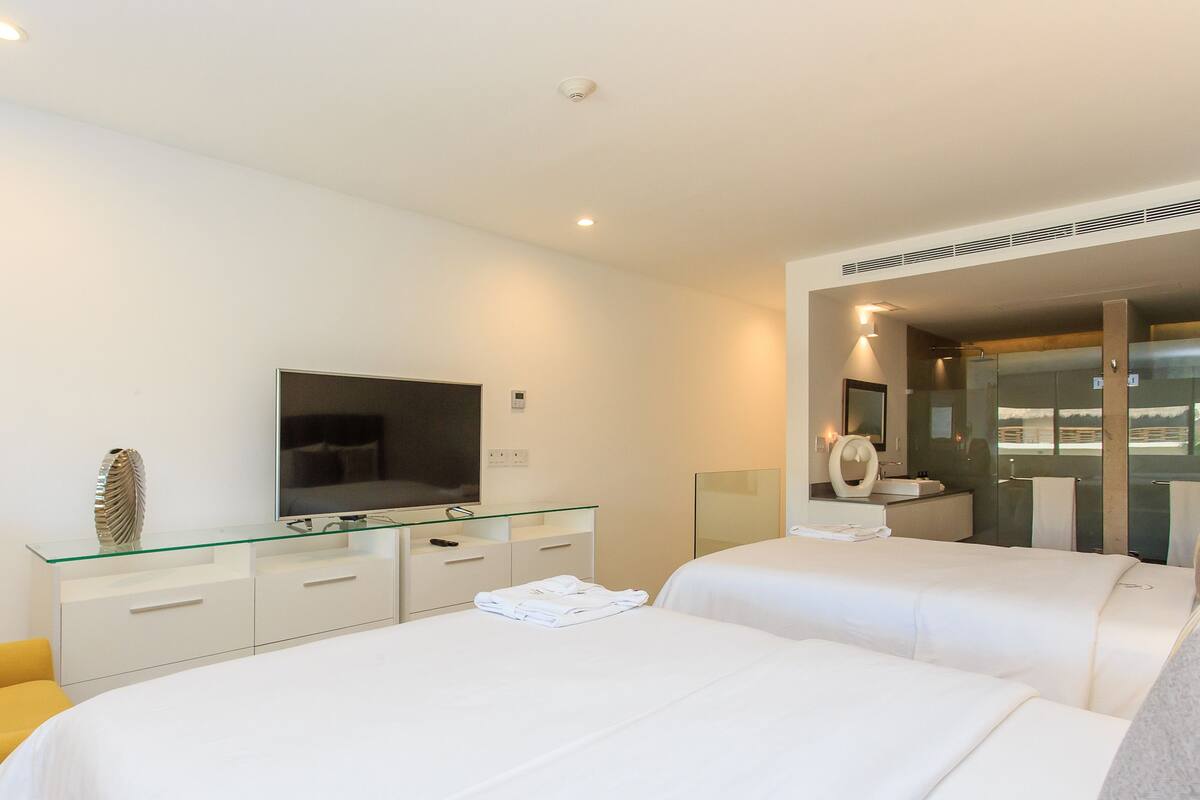 A modern hotel room with a flat-screen TV, a glass-topped desk, a bed with white linens, and a large mirror.