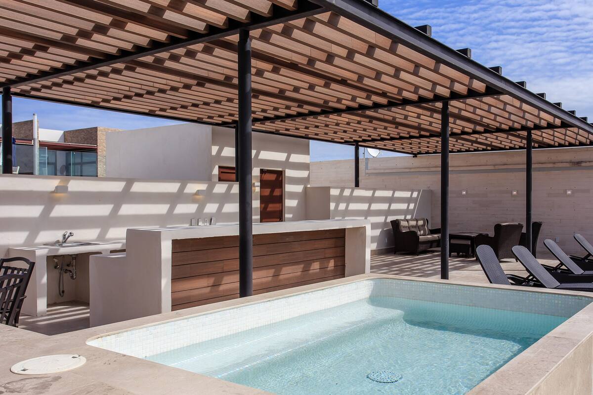 A rooftop pool area with a wooden pergola, lounge chairs, and a built-in seating area.