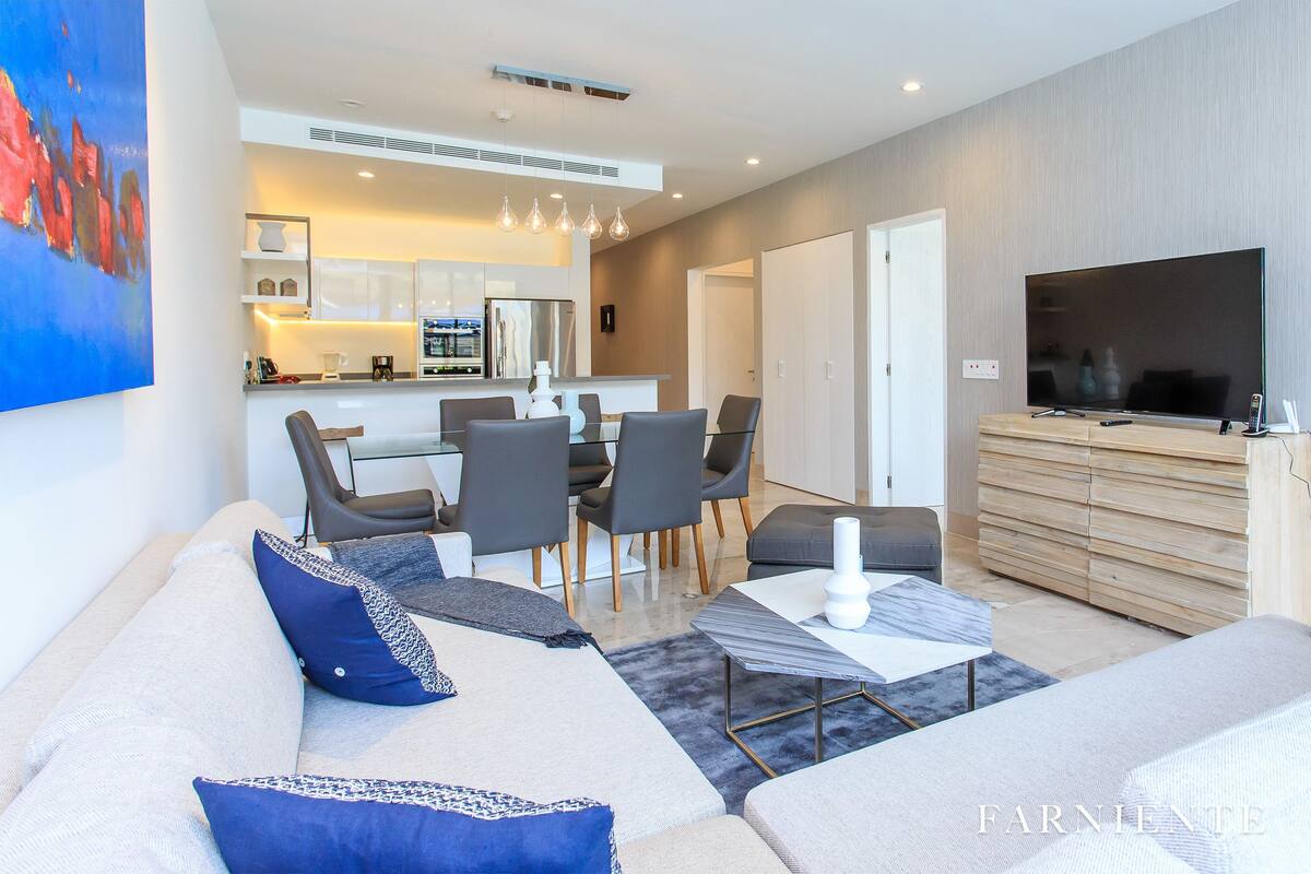 A modern living room with a sofa, a dining area with chairs, a TV, and a kitchen in the background.