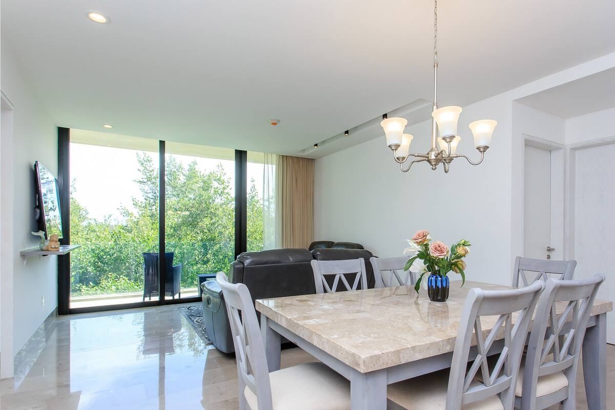 A modern dining area with a marble table, white chairs, a chandelier, and a view of greenery outside.