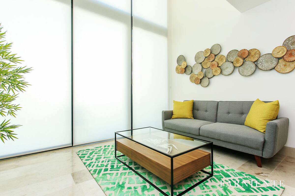 A modern living room with a glass coffee table, a grey sofa with yellow cushions, a patterned rug, and a wall art piece.