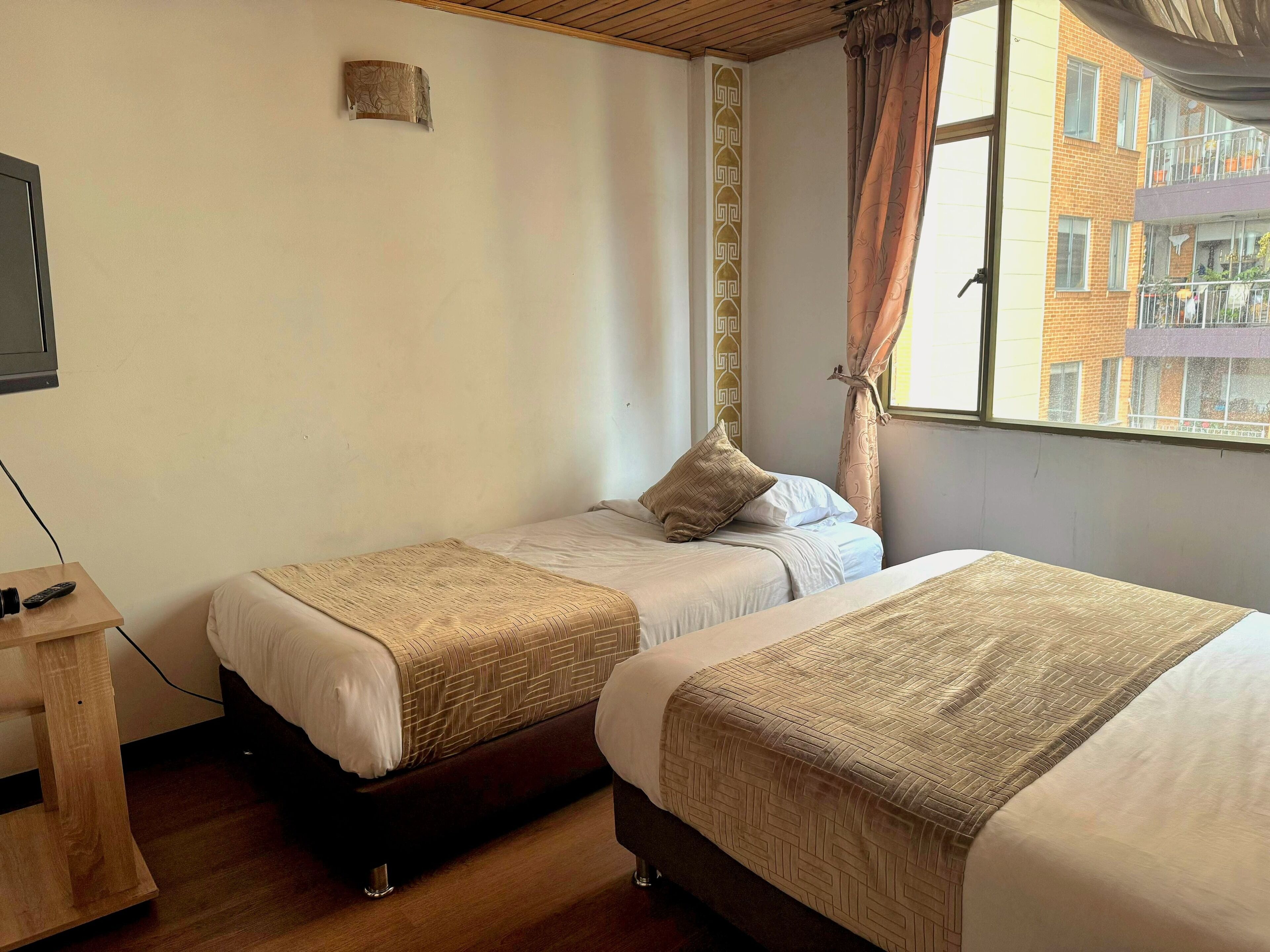 Een hotelkamer met twee enkele bedden, een houten nachtkastje, een televisie en een raam met gordijnen.