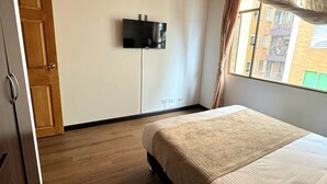 Un dormitorio con techo de madera, una cama con colcha color beige, un televisor en la pared y una ventana con cortinas.