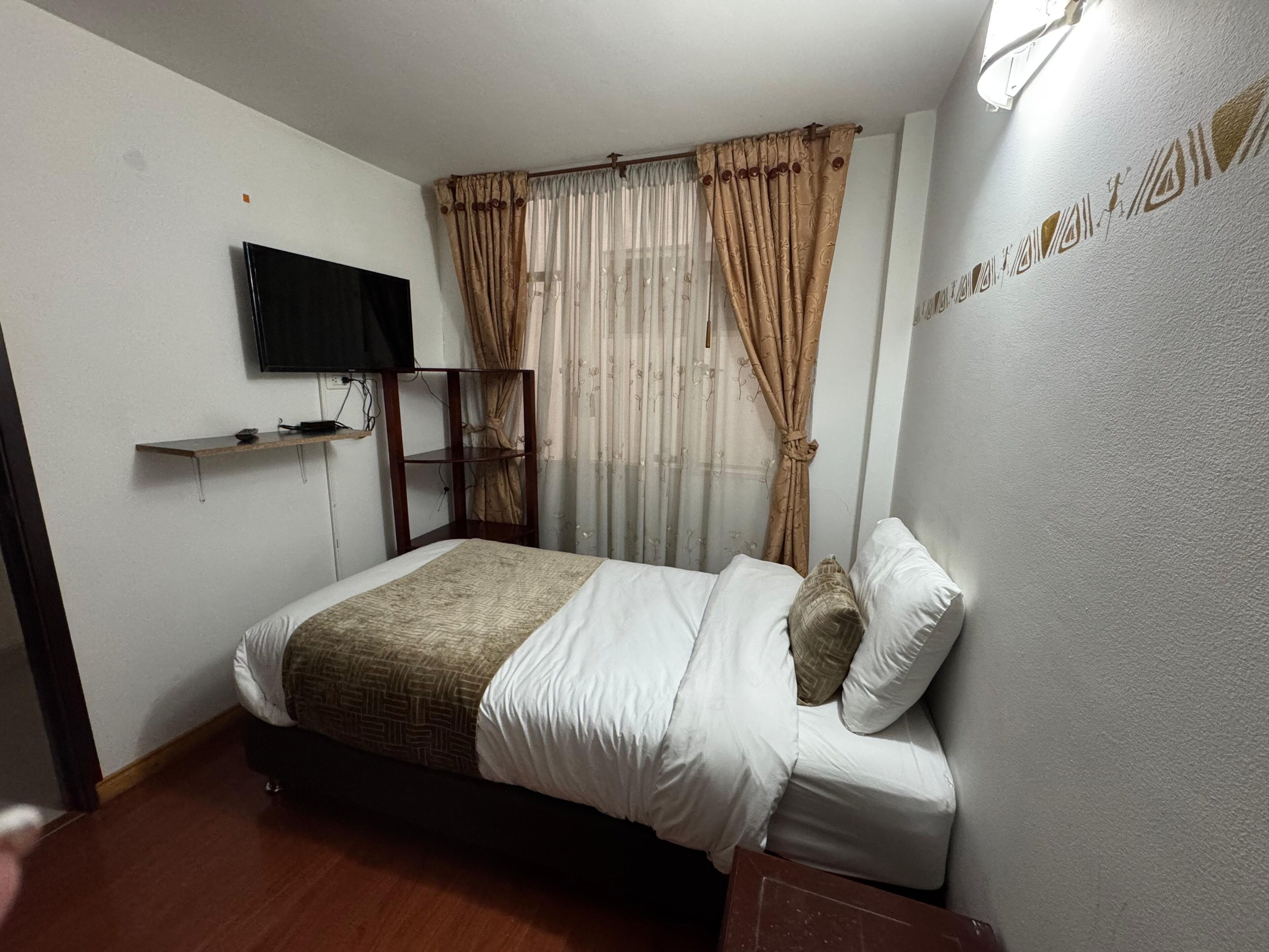 Une chambre d’hôtel avec un lit, un téléviseur fixé au mur et une fenêtre avec des rideaux.