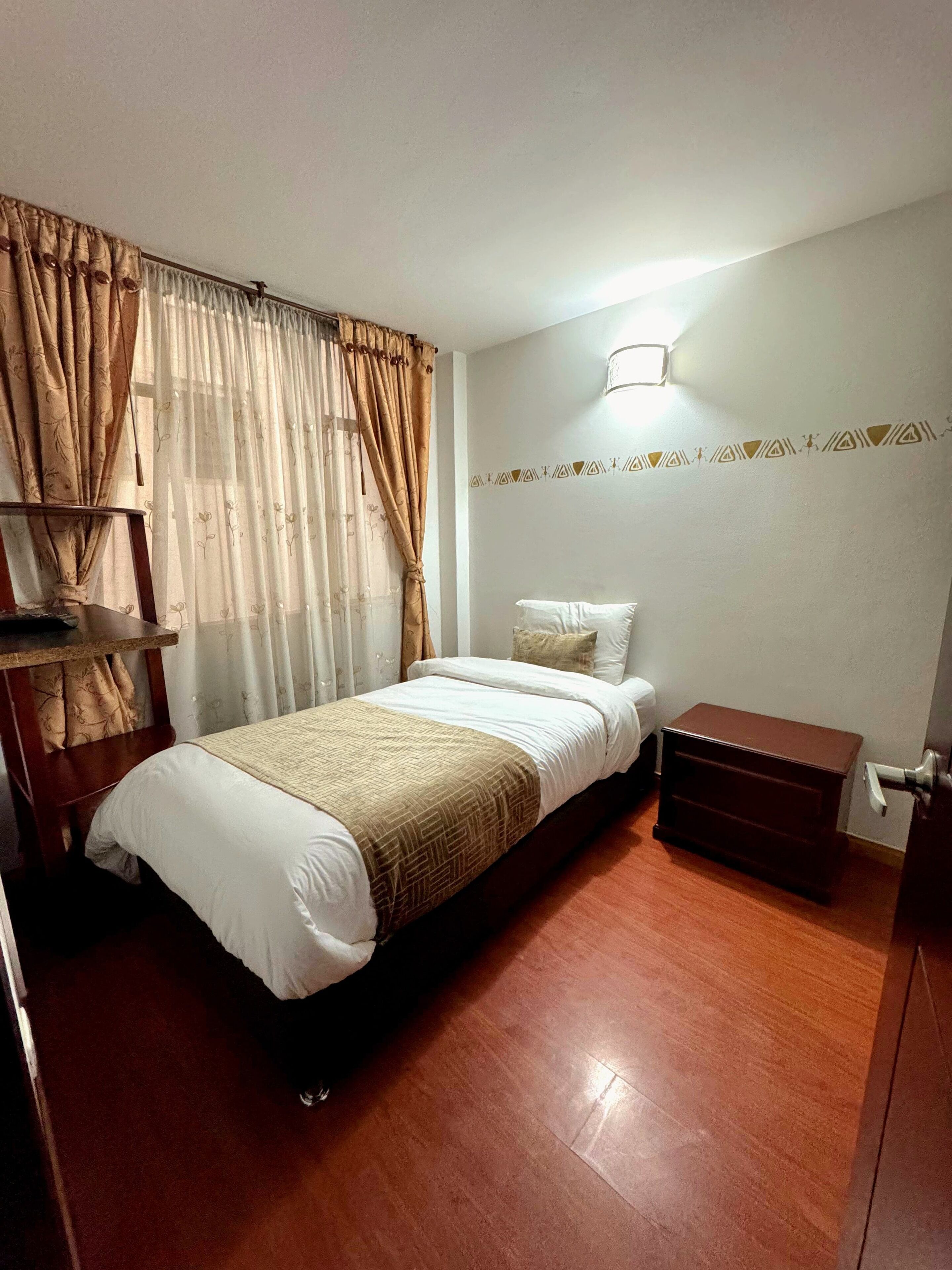 A bedroom with a bed, a bedside table, a wooden nightstand, a wall-mounted light, and a window with curtains.
