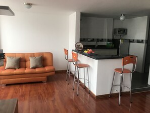 Un espacio habitable compacto con sofá, dos taburetes altos, una zona de cocina con microondas y una pequeña isla.