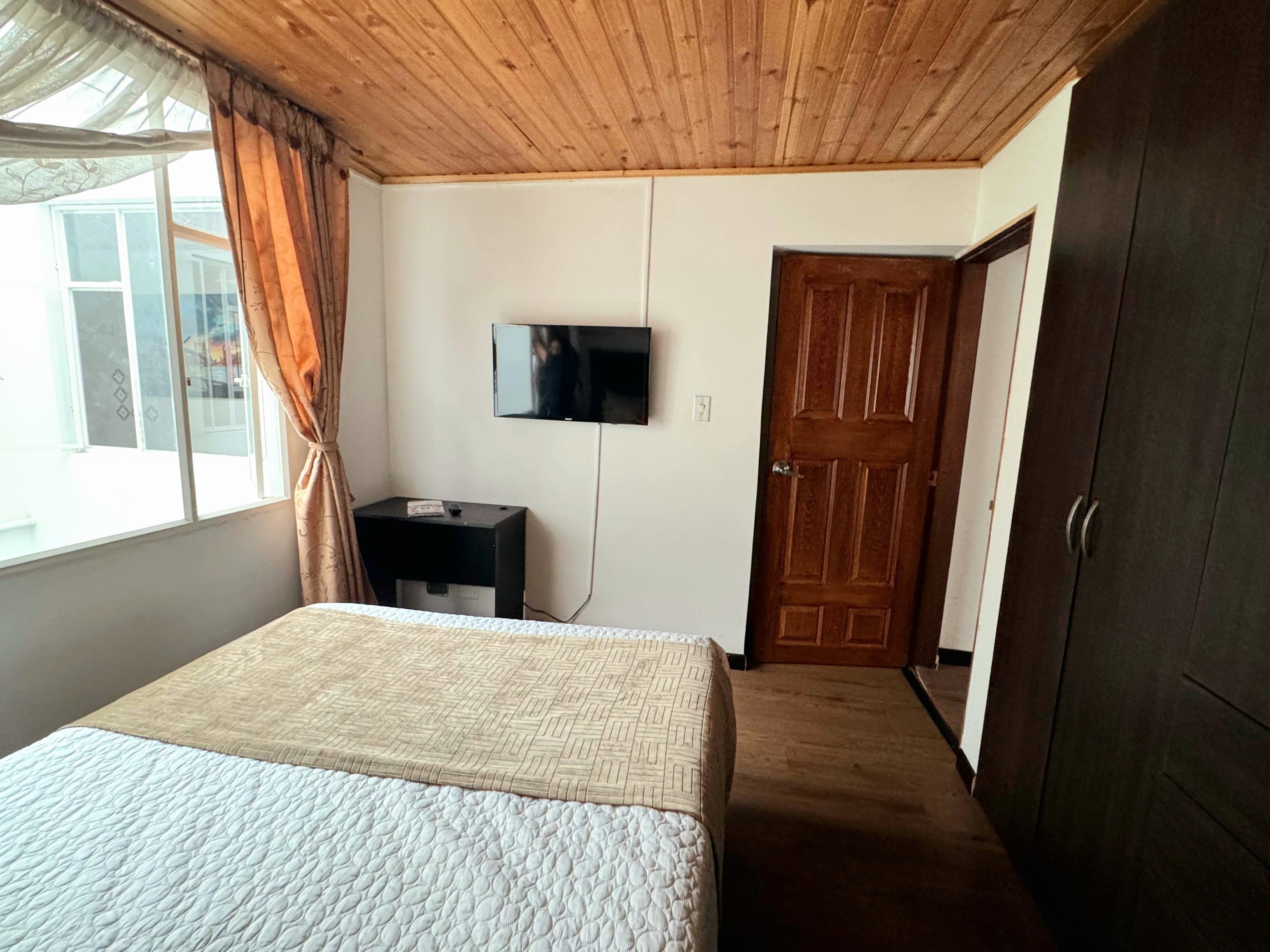 A bedroom with a bed, a TV mounted on the wall, a wardrobe, and a window with curtains.
