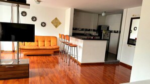 Una sala de estar moderna con sofá, televisor y una zona de cocina con refrigerador y microondas.