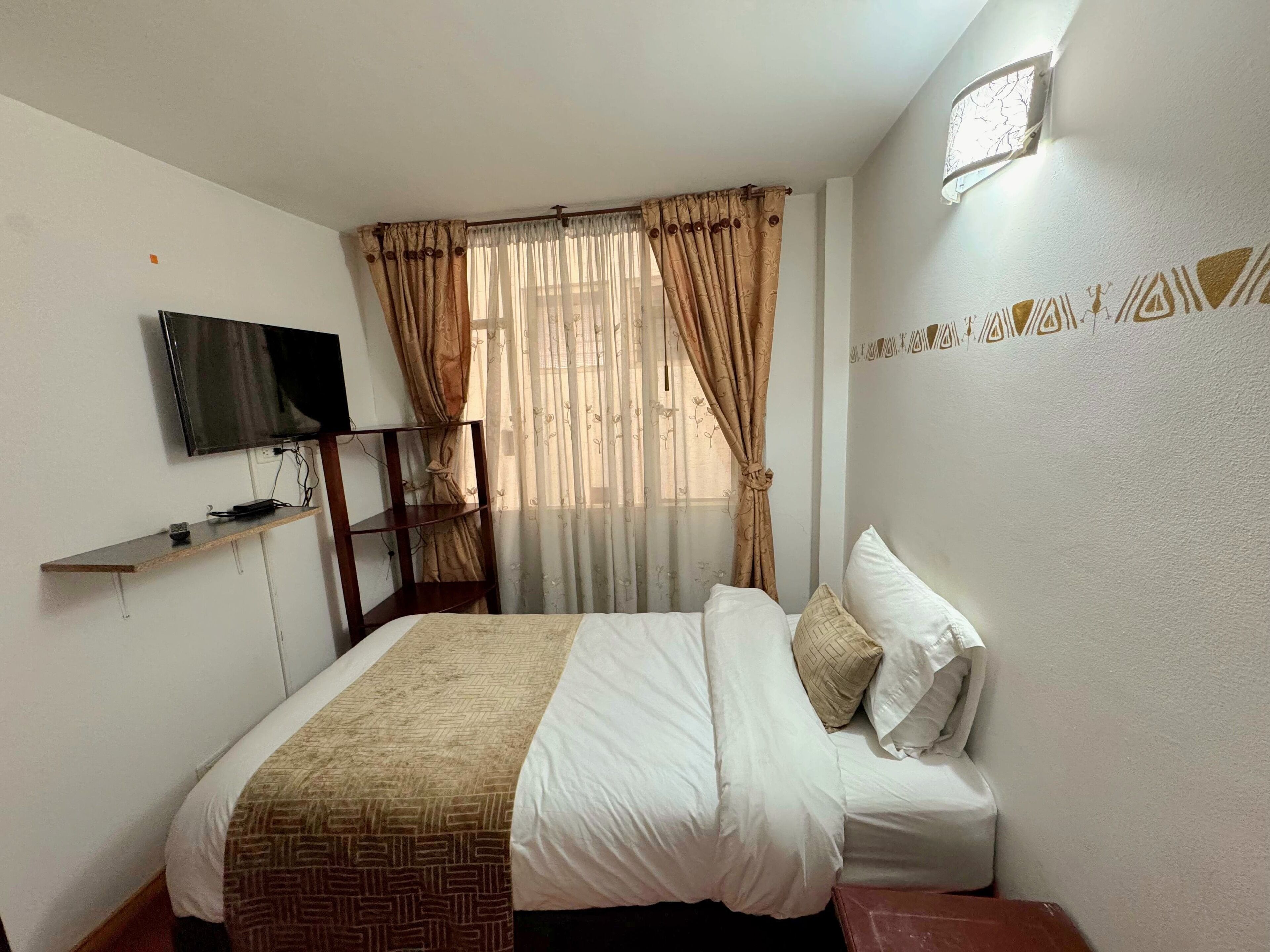 Un dormitorio con una cama, un televisor montado en la pared, un estante y una ventana con cortinas.