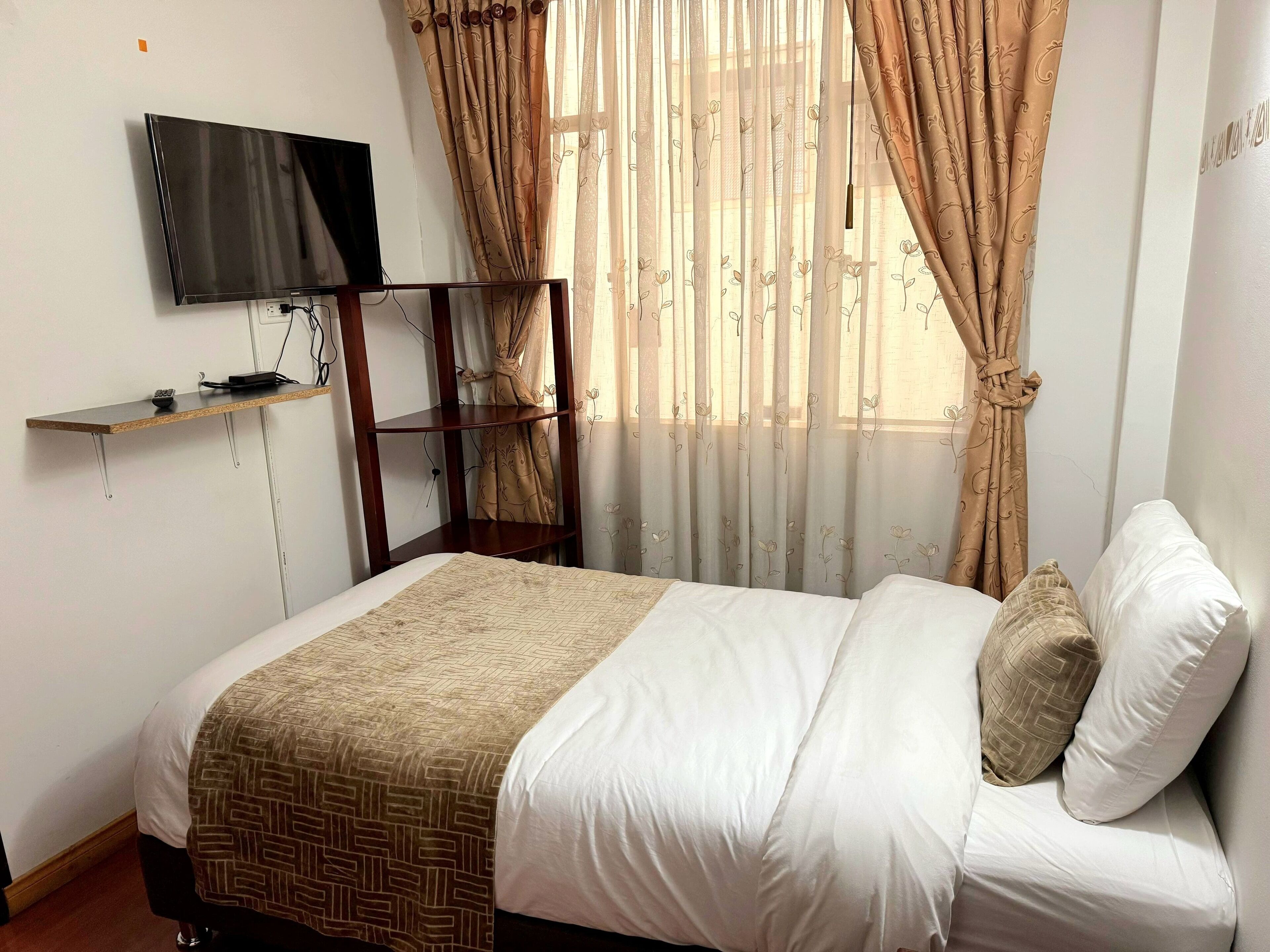 Un dormitorio con una cama, televisor montado en la pared, estante de madera y cortinas traslúcidas con motivos florales.