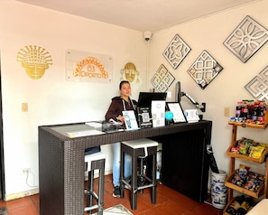 Un mostrador de recepción con una computadora, un teléfono y un terminal de pago. Se observan adornos en la pared y una estantería con aperitivos.