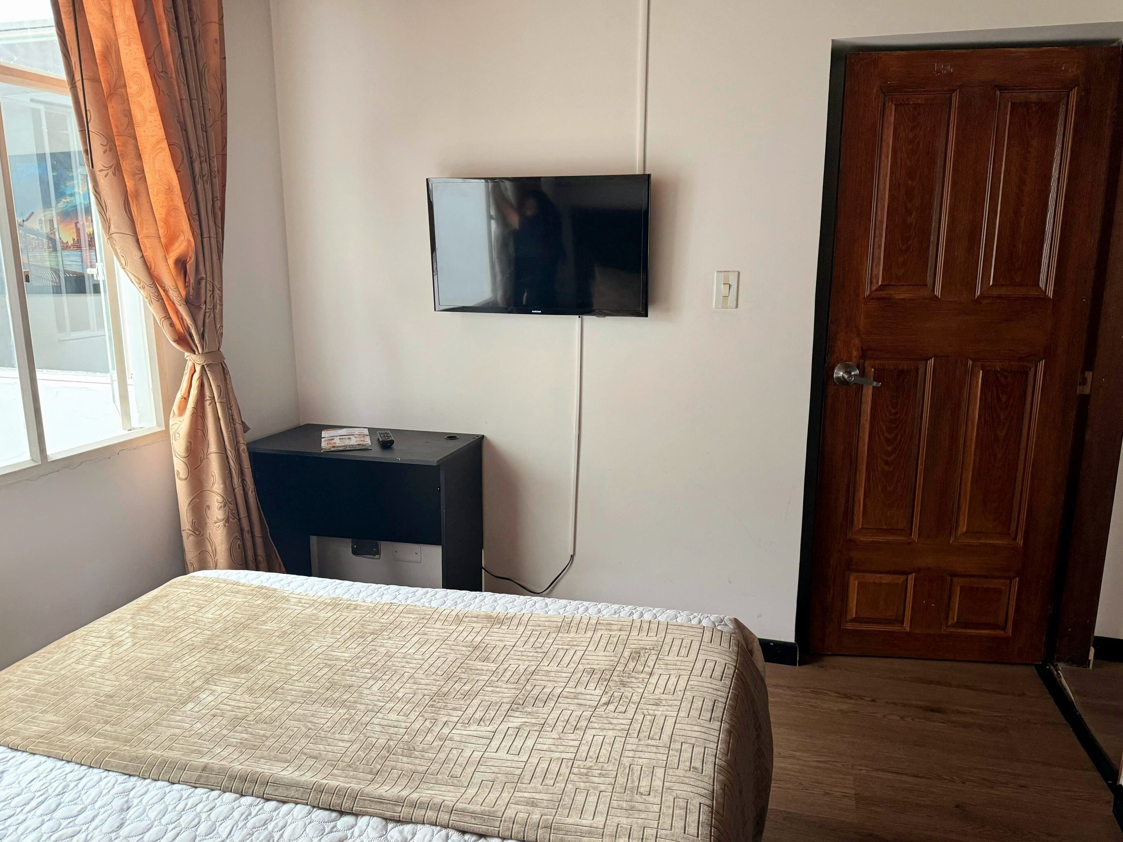 A bedroom with a bed, a TV mounted on the wall, a wooden door, and a window with curtains.