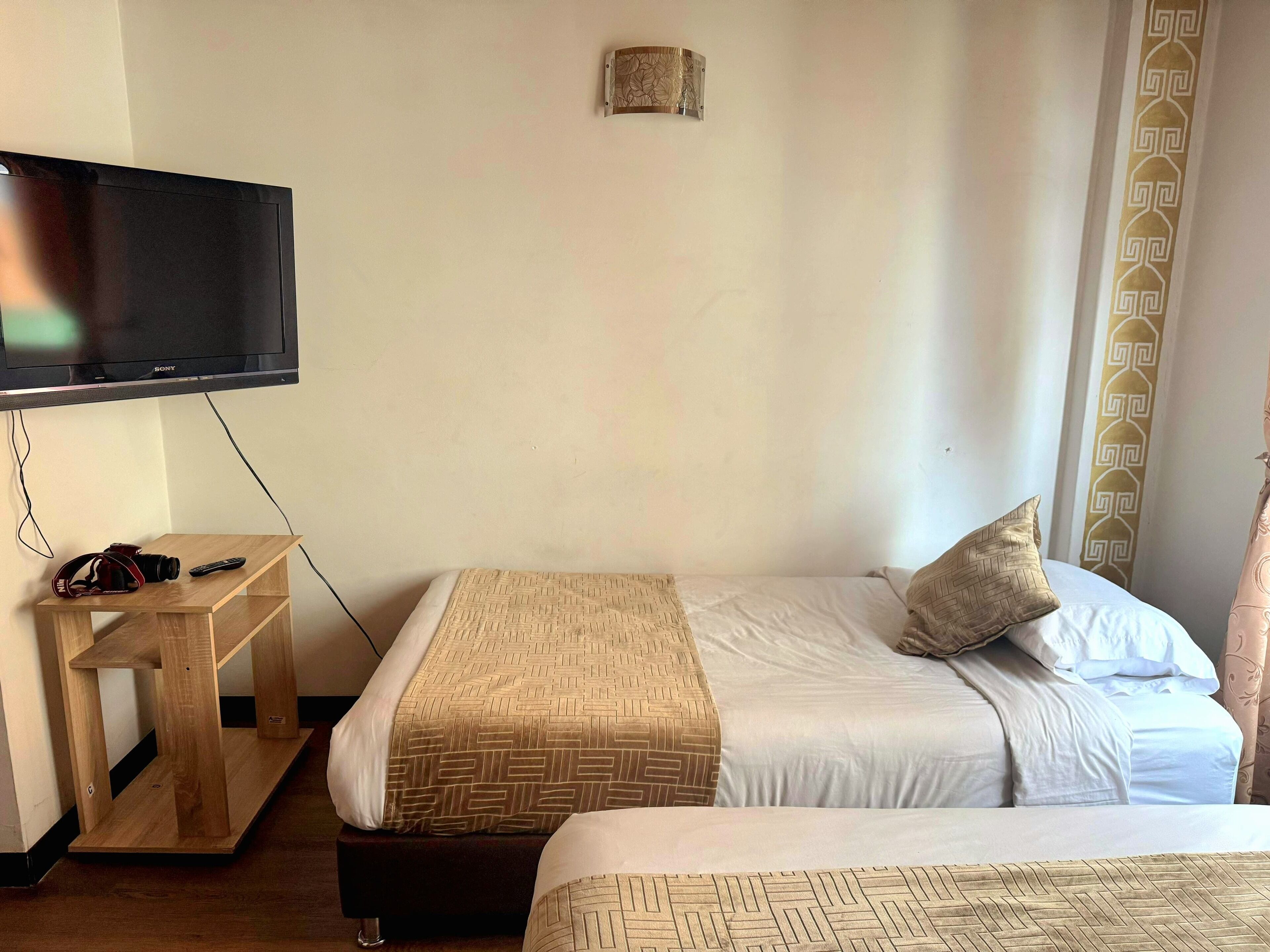 A hotel room with a bed, a TV mounted on the wall, a small wooden table, and a curtain with a geometric pattern.