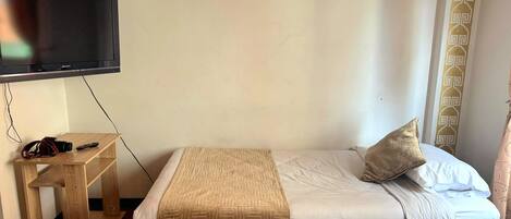 A hotel room with a bed, a TV mounted on the wall, a small wooden table, and a curtain with a geometric pattern.