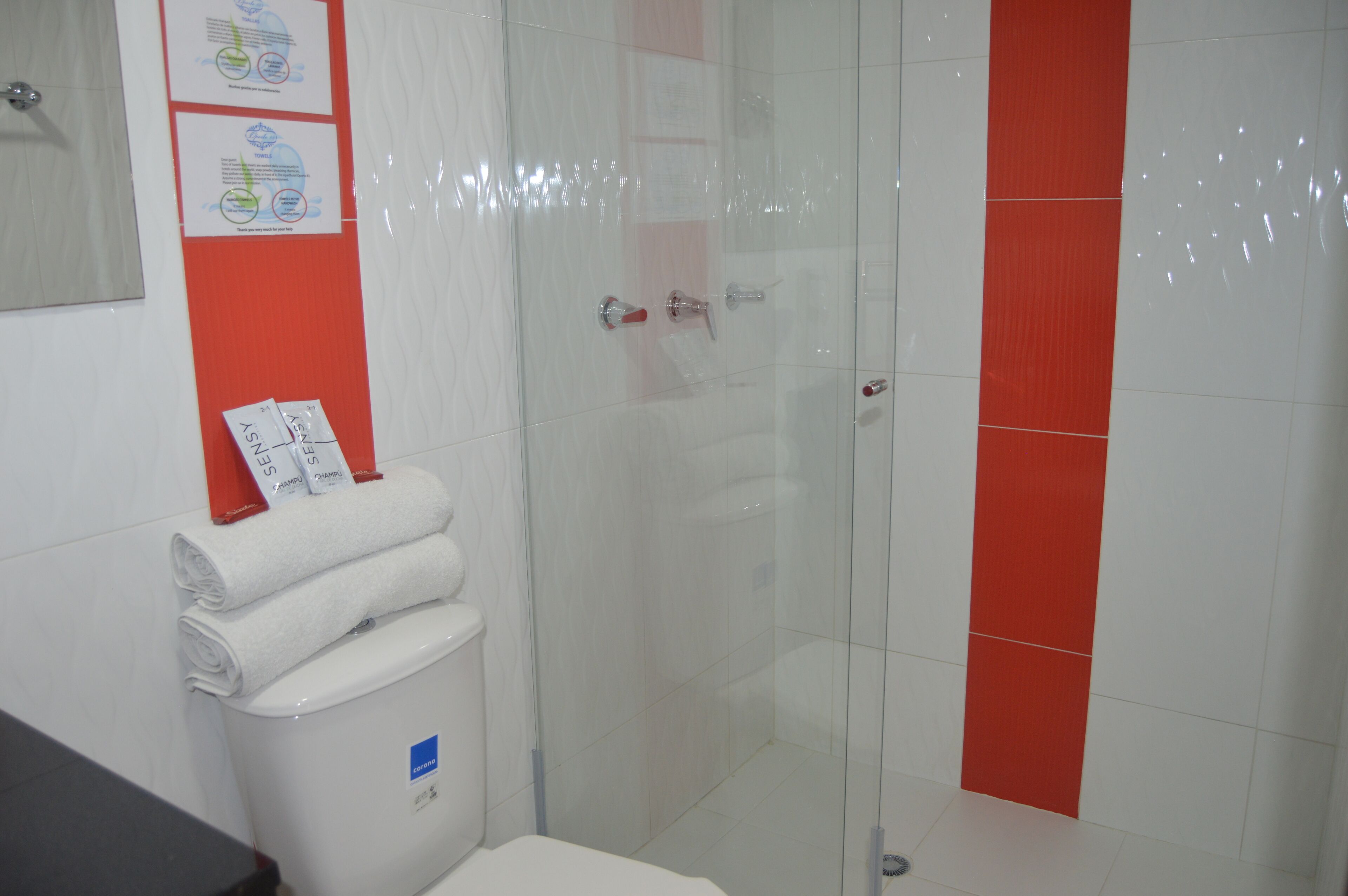 A bathroom with a glass shower enclosure, a toilet, and a sink. There are towels and a trash bin visible.