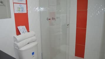 A bathroom with a glass shower enclosure, a toilet, and a sink. There are towels and a trash bin visible.
