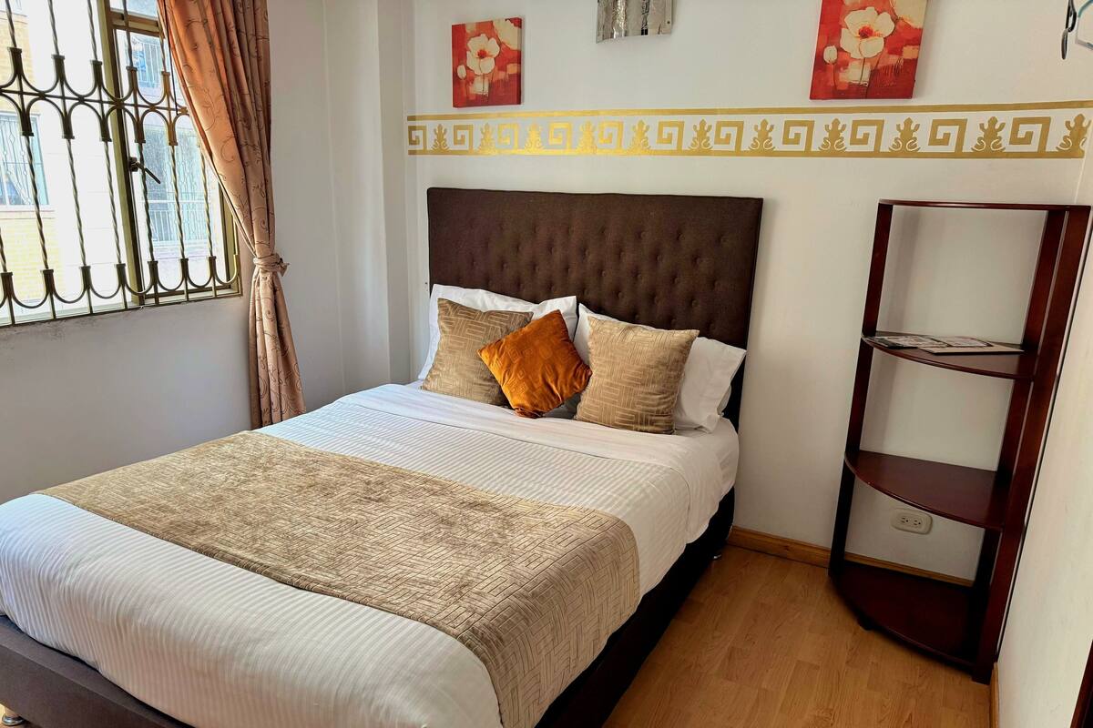 A neatly made bed with a patterned bedspread and pillows, a wooden floor, a window with metal bars, and a wall with decorative elements.